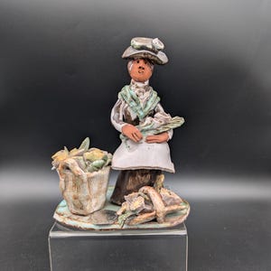 Peut inclure: Figurine en céramique d'une femme portant un chapeau et un tablier, tenant des fleurs. Un panier de fleurs et un panier de fleurs sont sur une base. La figurine est peinte dans des tons terre avec des accents de vert et de blanc.