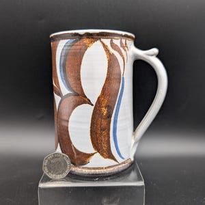 May include: A ceramic mug with a white base, adorned with brown and blue abstract patterns. The mug has a white handle and a brown rim. The mug is displayed on a clear acrylic stand against a black background.