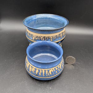 May include: Two handmade ceramic bowls. The bowls are blue with a beige band featuring a repeating pattern. The bowls are different sizes, with the larger bowl in the background and the smaller bowl in the foreground.