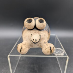 May include: A whimsical ceramic turtle figurine with large, expressive eyes and a smiling face. The turtle has a beige body with dark brown markings and is displayed on a clear acrylic stand. A coin rests on the stand.