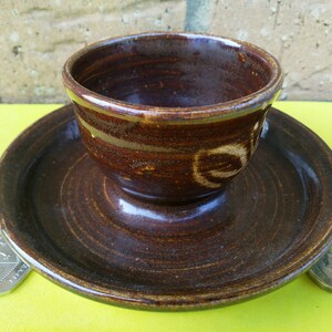 May include: A brown ceramic egg cup with a saucer. The cup and saucer have a swirl pattern and a gold rim.