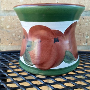 May include: A white ceramic vase with a green rim and base. The vase is decorated with a hand-painted design of red apples with green leaves.