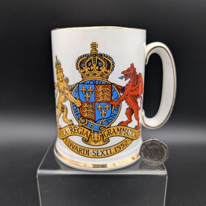 Può includere: Tazza in ceramica bianca con bordo e manico dorati, con un colorato stemma con corona, leoni e un drago. La tazza ha del testo attorno allo stemma ed è esposta su un supporto trasparente.