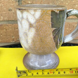 May include: A ceramic mug with a white, brown, and green glaze. The mug has a handle and a pedestal base. The mug is 7.6 cm tall.