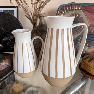 May include: Two ceramic pitchers with a matte white finish and vertical tan stripes. Each pitcher features a curved handle, a pouring spout, and a solid tan base. The design is consistent across both vessels, showcasing a clean, two-tone aesthetic.