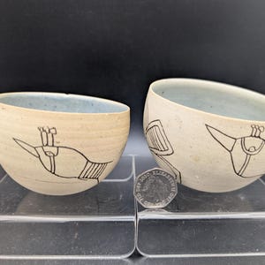 May include: Two round ceramic bowls with a light beige exterior and a pale blue interior. The exterior features black line drawings of stylized bird-like figures. A British fifty pence coin is placed between the bowls for scale reference.