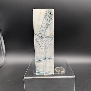 Könnte beinhalten: Eine rechteckige Keramikvase mit einem strukturierten, hellblauen und weißen Design. Die Vase weist eine Reihe von vertikalen und gebogenen Linien auf, die ein abstraktes Muster bilden. Eine Münze liegt auf dem transparenten Acrylständer.