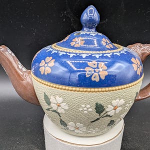 May include: A blue and white ceramic teapot with floral designs. The teapot has a brown handle and spout. The teapot is decorated with white flowers and green leaves on a light brown background.