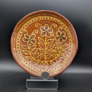 May include: A round, brown ceramic plate with a floral design. The plate features three stylized flowers with yellow and black accents, surrounded by a decorative border. The plate is displayed on a clear stand against a black background.