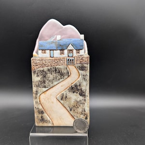 Può includere: Un'opera d'arte in ceramica raffigurante una casa con tetto blu e una strada tortuosa. La casa è posta su un muro di pietra, con una montagna rosa sullo sfondo. Il pezzo è esposto su un supporto trasparente.