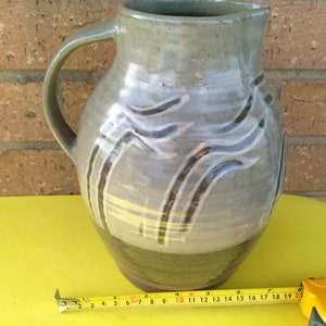 Puede incluir: Una jarra de cerámica gris con un asa marrón y un diseño abstracto en blanco y negro. La jarra está sobre una superficie amarilla.