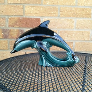 May include: A glossy ceramic dolphin figurine featuring a sleek black upper body and a deep teal lower section. The dolphin is depicted in a dynamic leaping pose, emerging from stylised, swirling teal waves that form the base of the sculpture.