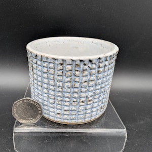 May include: A cylindrical ceramic planter featuring a textured grid pattern in light blue and dark brown tones. The interior is smooth and white. A 2020 British fifty pence coin is placed next to the planter to provide a clear sense of scale.