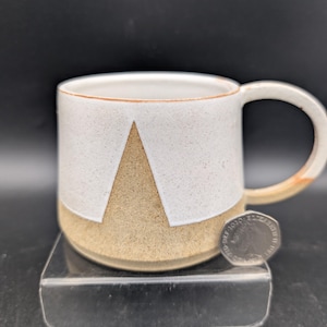 May include: A ceramic mug with a speckled white glaze on the upper portion and a natural tan clay base featuring a triangular cutout design. A British twenty pence coin is placed next to the mug for scale, showing the relative size of the vessel.