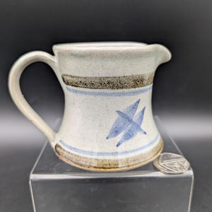 May include: A ceramic jug with a handle and spout. The jug is off-white with brown and blue painted stripes and a blue leaf design. The jug is sitting on a clear acrylic stand.