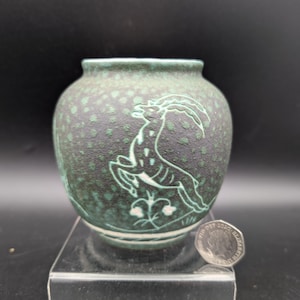 Può includere: Un piccolo vaso rotondo verde scuro con un disegno di cervo che salta in bianco. Il vaso ha una consistenza maculata e un bordo turchese. Una moneta è accanto al vaso per il confronto delle dimensioni.