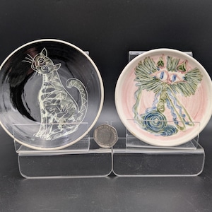 May include: Two decorative ceramic plates with cat designs. One plate is black with a white cat illustration, the other is pink with a colorful cat design. The plates are displayed on clear stands.