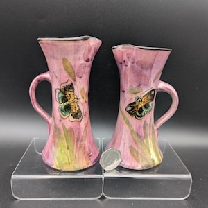 May include: Two pink ceramic vases with handles, each adorned with a butterfly design. The vases have a flared shape and are decorated with green and yellow accents. The vases are displayed on a clear acrylic shelf.