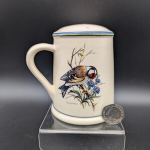 Peut inclure: Mug en céramique crème avec une anse et un couvercle. La tasse présente une illustration détaillée d'un chardonneret perché sur une branche avec des fleurs bleues. Le mot "Goldfinch" est imprimé en dessous.