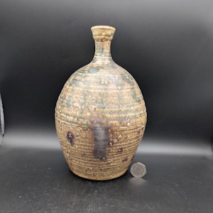 May include: A round, textured ceramic bottle with a narrow neck, featuring a mottled brown, tan, and dark green glaze with horizontal ridges. A silver-coloured coin is placed at the base for scale, providing a clear reference for the object's size.