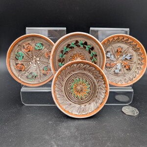 May include: Four small, round, hand-painted ceramic plates with intricate designs. The plates feature a brown rim and a central design with orange, green, and blue accents. The plates are displayed on a clear stand against a black background.