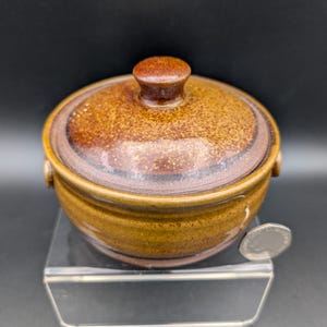 Op de afbeelding: Een kleine, ronde keramische schaal met deksel in bruine tinten. De schaal heeft een gespikkelde textuur en een klein handvat op het deksel. De schaal staat op een helder, schuin oppervlak.