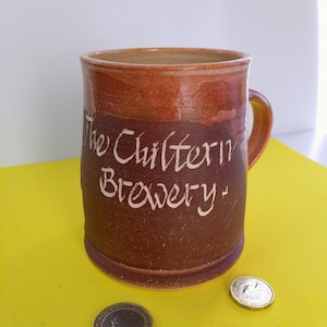 Könnte beinhalten: Eine braune Keramiktasse mit der Aufschrift "The Chiltern Brewery". Zwei britische Münzen liegen auf einer gelben Oberfläche neben der Tasse.