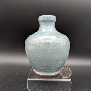 Może przedstawiać: Mały, ręcznie robiony ceramiczny wazon o zaokrąglonym korpusie i wąskiej szyi. Wazon jest szkliwiony w odcieniach niebieskiego i szarego, z subtelnym nakrapianiem. Podstawa ma czerwonobrązową obwódkę. Wazon jest eksponowany na przezroczystym, akrylowym stojaku.