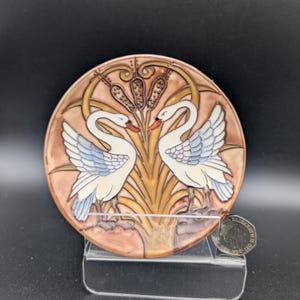 Könnte beinhalten: Eine runde, dekorative Keramikplatte mit zwei weißen Schwänen, die sich gegenüberstehen, mit blauen und grauen Flügeldarstellungen. Die Platte hat einen braunen und beigen Hintergrund mit stilisierten Rohrkolben und Blättern.