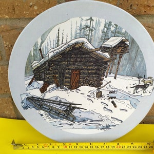 May include: A blue ceramic plate featuring a winter scene illustration. The scene shows a log cabin with a snowy roof, surrounded by trees and snow. Two dogs are walking in the snow. The plate has a white border.