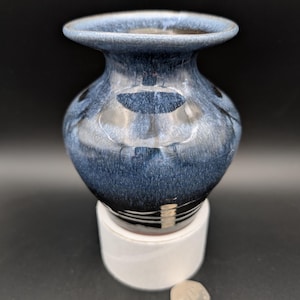 Puede incluir: Un pequeño jarrón de cerámica azul oscuro con un acabado brillante. El jarrón tiene un cuerpo redondeado y un borde acampanado.