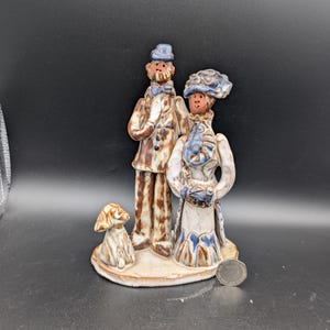 May include: A ceramic figurine depicting a couple and a small dog standing on a circular base. The figures feature brown and white glazed surfaces with blue accents on their hats and clothing. A silver coin is placed next to the base for scale.