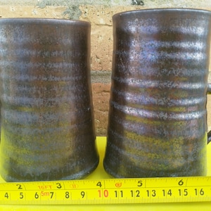 Könnte beinhalten: Zwei Keramikbecher mit dunkelbrauner Glasur und einem metallischen Schimmer. Die Becher sind etwa 10 Zoll hoch und haben eine abgerundete Form. Die Becher stehen auf einer gelben Oberfläche mit einem Maßband.