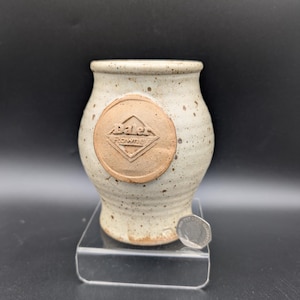 May include: A small, speckled ceramic jar with a wide mouth and a rounded body. The jar is off-white with brown speckles and a circular, raised emblem that reads "Dale Rowney". The jar sits on a clear acrylic stand.