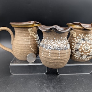 May include: Three brown ceramic mugs and vases with decorative white floral patterns. The mug on the left has a handle. The other two have ruffled rims. The pottery is on a clear acrylic stand.