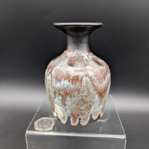 May include: A small, handcrafted ceramic vase with a dark brown rim and a body featuring a blend of brown, blue, and cream colors. The vase has a unique, textured appearance and a flared top.