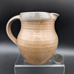 Puede incluir: Una jarra de cerámica hecha a mano con un cuerpo redondeado y un asa curva. La jarra es de un marrón cálido y terroso con un borde más claro y moteado. Tiene un pico para verter fácilmente. Una moneda está al lado de la jarra.