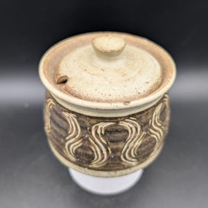 Op de afbeelding: Een kleine, witte suikerpot van keramiek met een deksel. De pot heeft een bruin en wit swirl-ontwerp.