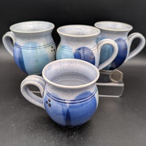Könnte beinhalten: Vier Keramikbecher mit blau-weißer Glasur. Jeder Becher hat einen abgerundeten Körper, einen Henkel und ein Design aus blauen Kreisen und vertikalen Streifen. Die Becher sind handgefertigt und haben eine rustikale, handwerkliche Ausstrahlung.