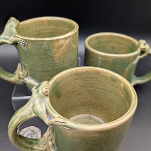Könnte beinhalten: Drei handgefertigte Keramikbecher in verschiedenen Größen. Die Becher sind in Grün- und Brauntönen glasiert und haben einzigartige, tintenfischförmige Griffe. Das Innere der Becher ist hellgrün. Die Becher werden auf einer transparenten Oberfläche vor dunklem Hintergrund präsentiert.