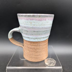 May include: A handcrafted ceramic mug with a brown base and a light blue and pink striped top. The mug has a curved handle and is displayed on a clear acrylic surface against a black background. The mug is approximately 10 cm tall.