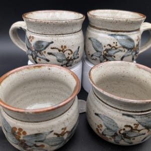 May include: Four ceramic mugs with handles. The mugs are beige with a speckled finish and feature a floral design in blue, brown, and black. The rims and handles are a lighter beige colour. The mugs are arranged in a group.