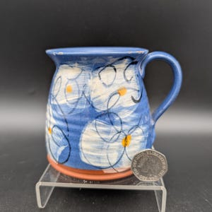 May include: A blue ceramic mug with white floral designs and orange centers. The mug has a handle and sits on a clear stand. The mug's rim and handle are a darker shade of blue.