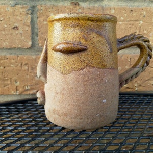 May include: A brown and tan ceramic mug with a face sculpted on the front. The mug has a twisted rope handle.
