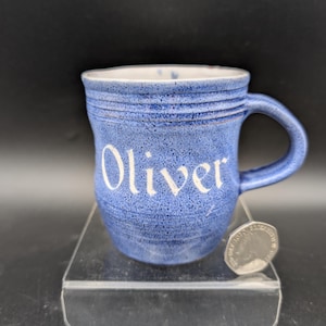 Może przedstawiać: Niebieski kubek ceramiczny z nakrapianym wykończeniem i napisem Oliver w białym kolorze z przodu. Kubek posiada poziome żłobienia przy krawędzi oraz zakrzywione ucho. Obok kubka umieszczono monetę dla porównania skali wielkości przedmiotu.