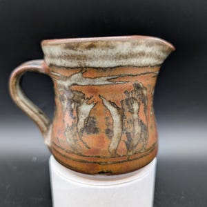 May include: A small brown and white ceramic pitcher with a handle. The pitcher has a design of stylized figures in brown, white, and orange.