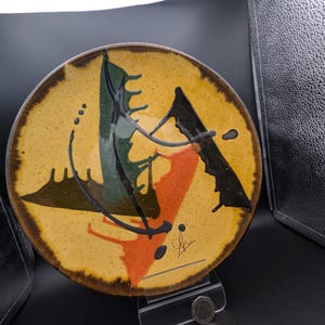 May include: A round ceramic plate with a yellow background and abstract art in green, orange, and black. The plate has a brown rim and a signature in the lower right corner. The plate is displayed on a clear stand.
