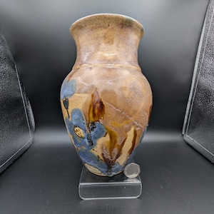 Könnte beinhalten: Eine handgefertigte Keramikvase mit breitem Körper und schmalem Hals. Die Vase weist eine Mischung aus braunen, beigen und blauen Farbtönen mit blütenartigen Mustern auf. Die Vase wird auf einem transparenten Ständer ausgestellt.