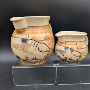 May include: Two ceramic pitchers, one large and one small, with a cream-colored rim and handle. The bodies are tan with a grid pattern and a fish design. The pitchers are on a clear acrylic surface.