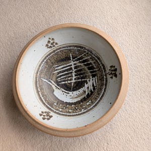 May include: A round ceramic bowl with a beige rim and a white interior. The bowl's centre features a dark brown design with white linear and abstract shapes. Small brown clusters are placed around the rim.